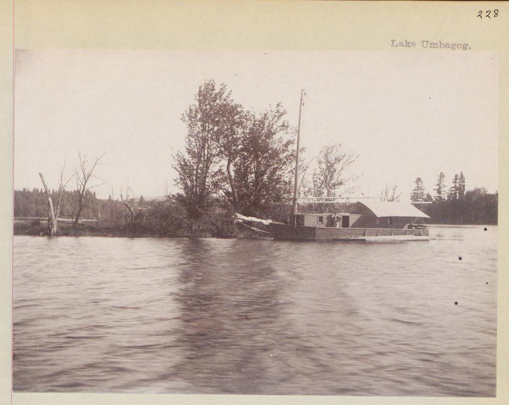 Black and white photograph of a small house boat on a lake with a person standing on deck.