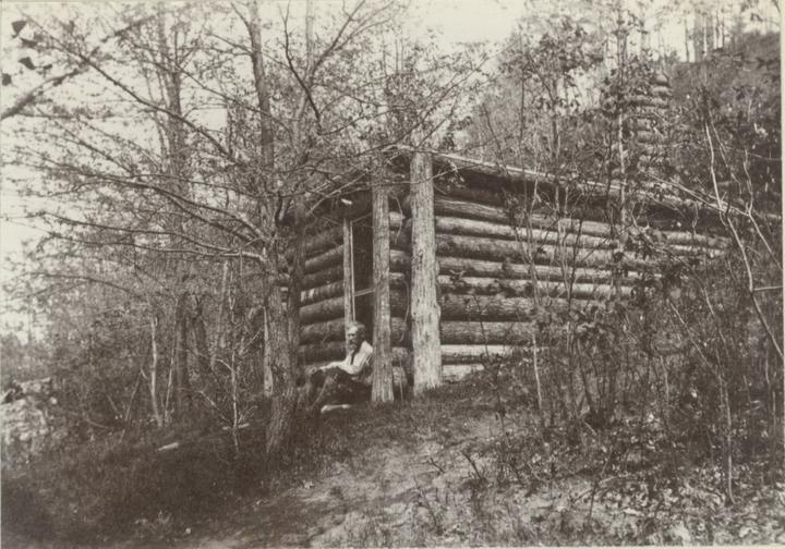 Brewster seated in front of cabin.